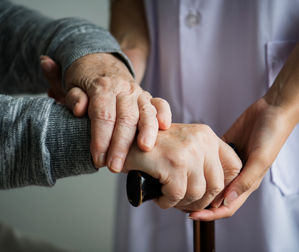 Pflegende Hände stützen eine ältere Person mit Gehstock – Symbol für Unterstützung, Vertrauen und Geborgenheit im Alter.
